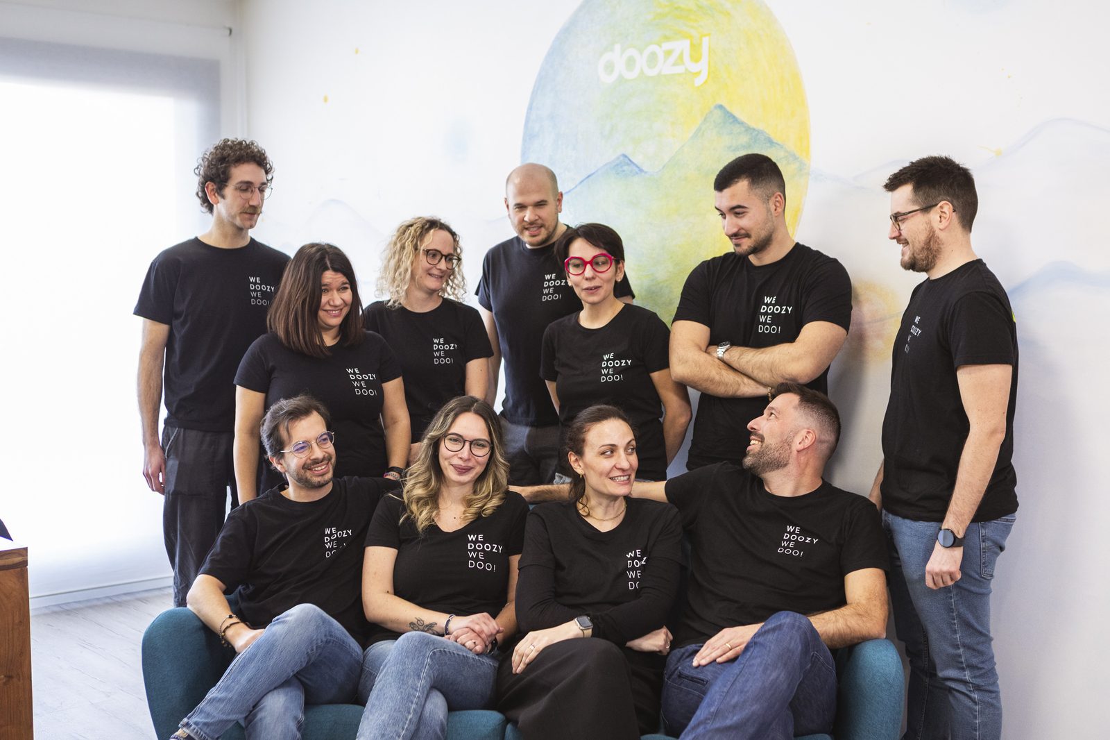 Il team Doozy al completo, dieci persone tra sedute sul divano e in piedi, davanti al murales del logo dipinto a mano nell'ufficio di Selvazzano